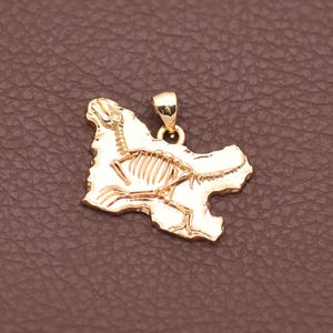 May include: A gold-toned pendant featuring an irregular, rock-like shape with an engraved skeleton of a four-legged animal. The pendant includes a small, polished bail at the top for attachment to a chain, showcasing a metallic finish.