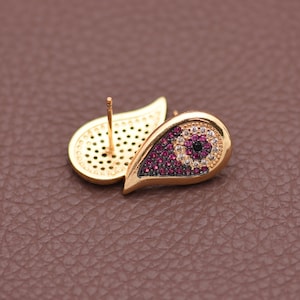 May include: Gold-toned stud earrings with a teardrop shape. The earrings feature a central design of small, round, dark red and white gemstones, with a black center. The earrings are set against a brown background.