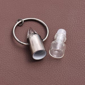 May include: A silver-colored bullet-shaped essential oil bottle with a clear crystal-like top and a small, clear glass bottle. The bottle has a silver ring attached. The interior of the bullet-shaped bottle is white.