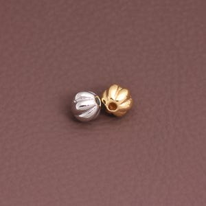 Puede incluir: Dos cuentas decorativas. Una es plateada, la otra dorada. Ambas tienen un diseño acanalado en forma de calabaza. Las cuentas son pequeñas, probablemente utilizadas para la fabricación de joyas o manualidades.