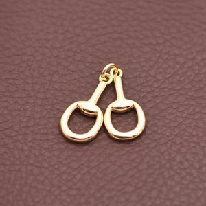 May include: Two gold-colored horse bit charms. Each charm features a rounded oval shape connected to a straight bar with a small loop at the top. The charms are displayed against a brown background.