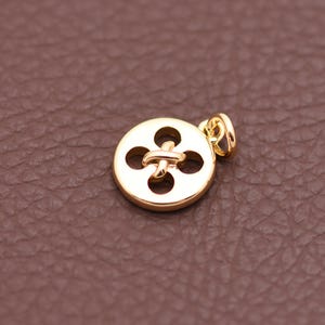 May include: A gold-colored button pendant with a four-petal design and a loop for a chain. The pendant is round and has a shiny finish. The background is a textured brown surface.