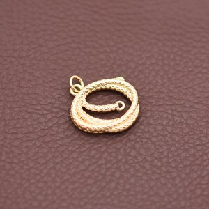 Op de afbeelding: Een goudkleurige kettinghanger met een textuur, touwachtig ontwerp. De hanger is opgerold in een cirkelvorm met een kleine lus aan de bovenkant om aan een ketting te bevestigen. De achtergrond is effen bruin.