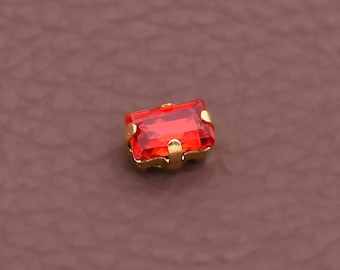 May include: A rectangular, faceted, red gemstone set in a gold-colored metal base. The gemstone is the focal point, with a vibrant, transparent appearance. The setting has four prongs holding the stone.