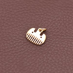 May include: A gold-coloured comb-shaped pendant with a small loop for a chain. The pendant has a rounded top with two small holes and vertical teeth. The background is a textured brown surface.