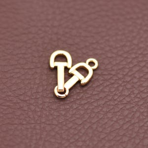 Puede incluir: Dos colgantes metálicos dorados con forma de bocado de caballo. Un colgante está en posición vertical y el otro está inclinado hacia la derecha. Los colgantes tienen un acabado brillante sobre un fondo marrón.