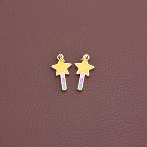 May include: Two gold-colored star-shaped charms with yellow centers and pastel-colored wands. The charms have a loop at the top for hanging. The background is a solid, dark brown color.