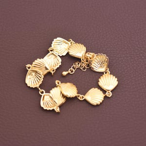 May include: A gold-colored bracelet featuring seashell charms. The bracelet has multiple seashell-shaped links connected by small rings and a chain for adjusting the size. The bracelet is displayed on a brown surface.