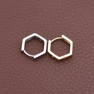 May include: Two hexagonal hoop earrings, one silver-toned and one gold-toned, are displayed against a brown background. The earrings have a geometric design and a hinged closure. The gold earring has a small, decorative element.