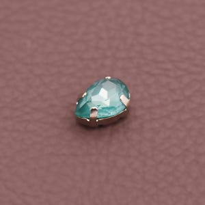 Può includere: Una gemma sfaccettata a forma di goccia, color turchese, con una montatura in metallo color argento. La gemma è posta su uno sfondo marrone. La gemma ha una superficie lucida e riflettente.