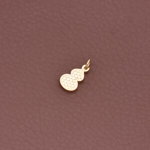 May include: A gold-coloured charm in the shape of a gourd. The charm has a textured surface with small, evenly spaced dots. It features a small loop at the top for attachment to a necklace or bracelet. The charm is set against a brown background.