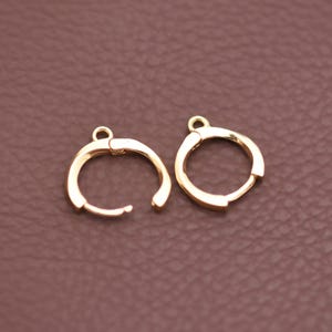 May include: Two gold-colored hoop earrings. Each earring has a circular design with a hinged closure. The earrings are displayed on a brown surface. The earrings are made of a shiny, metallic material.