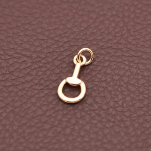 May include: A small, gold-colored charm with a ring at the top and a rounded, D-shaped bottom. The charm has a simple, elegant design, likely for jewelry making or crafting. The charm is shown against a brown background.
