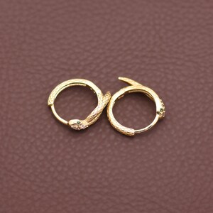 Puede incluir: Un par de pendientes de aro dorados. Cada pendiente presenta un diseño de serpiente con un cuerpo texturizado y una pequeña gema verde. Los pendientes se muestran sobre una superficie marrón.
