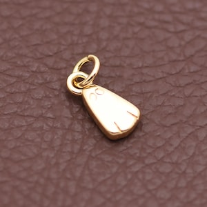 May include: A gold-coloured ghost-shaped charm. It features a loop for attachment and two small eye details. The ghost has two vertical lines at the bottom. The charm is likely for a necklace or bracelet.