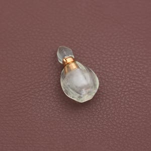 May include: A small, clear glass perfume bottle with a gold-colored band and a rounded, teardrop shape. The stopper is also clear and has a rounded, elongated shape. The bottle is set against a brown background.