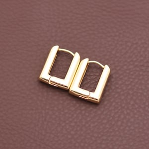 Könnte beinhalten: Ein Paar goldfarbene, rechteckige Creolen. Die Ohrringe haben eine polierte Oberfläche und ein schlichtes, modernes Design. Die Ohrringe werden auf einer braunen Oberfläche präsentiert.