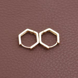 May include: A pair of gold-coloured hexagonal hoop earrings. The earrings have a geometric design and a hinged closure. The earrings are displayed on a brown surface.