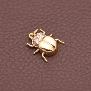 Peut inclure: Un charm doré en forme de scarabée avec une finition polie. Le bijou présente un design détaillé avec six pattes et une tête ornée de petites pierres précieuses claires. Le scarabée est posé sur un fond marron.