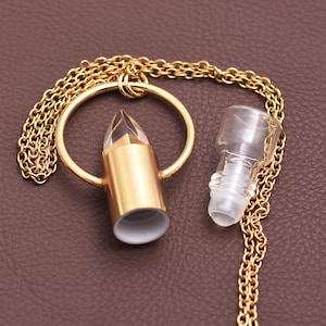 Può includere: Un ciondolo a forma di bottiglia di profumo in metallo color oro con una parte superiore in cristallo trasparente e sfaccettato e una bottiglia di vetro trasparente abbinata con un tappo a vite. Entrambi sono collegati da una catena d'oro. Il ciondolo è su una superficie marrone.