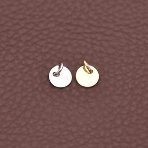 May include: Two small, round metal charms. One is silver-colored, the other is gold-colored. Each charm has a small loop at the top for attaching to a chain or bracelet. The charms are displayed on a brown leather-textured surface.
