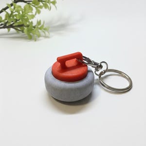 May include: A miniature curling stone keychain. The keychain features a gray stone with a red handle. The stone is attached to a silver-colored keyring and clasp. The background is white with a sprig of green leaves.