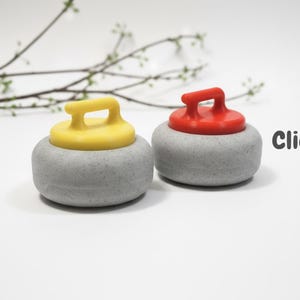 May include: Two miniature curling stones, one with a yellow handle and the other with a red handle, sit side-by-side. The stones are gray with a rounded shape. The word "Clicky!" is written in black. A sprig of greenery is in the background.
