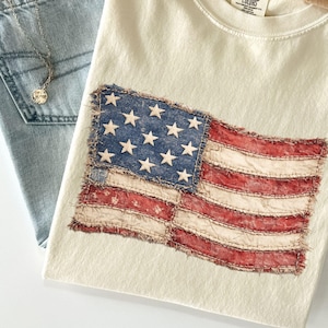 May include: Cream-colored t-shirt featuring a distressed American flag design. The flag is stitched onto the shirt with red, white, and blue colors. The shirt is folded and placed with a pair of blue jeans and a tan hat.
