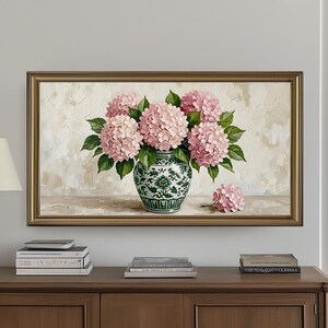 May include: A framed painting of pink hydrangeas in a green and white vase. The painting is displayed above a dark brown wooden cabinet with three doors. A gold lamp and books are on the cabinet.