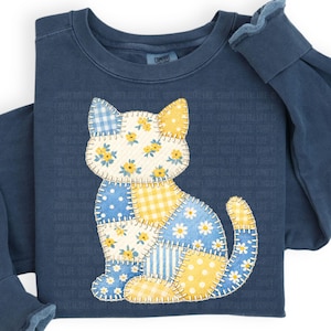 Könnte beinhalten: Marineblaues Sweatshirt mit einem Patchwork-Katzen-Design. Die Katze besteht aus verschiedenen Stoffmustern, darunter blaues Vichy, Blumen und Punkte. Das Sweatshirt hat lange Ärmel und einen Rundhalsausschnitt.
