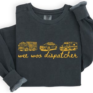 May include: A charcoal gray long-sleeve sweatshirt featuring a yellow graphic print of a fire truck, a police car, and an ambulance. Below the vehicles, the text "wee woo dispatcher" is printed in a yellow cursive font. The garment has a crew neckline.
