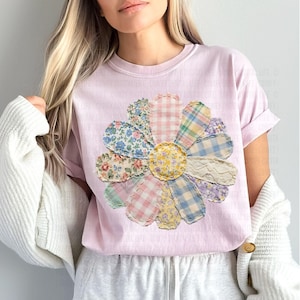 May include: A light pink t-shirt featuring a large, colorful flower design. The flower is made of various patterned fabrics, including floral, gingham, and lace, creating a patchwork effect. The shirt has short sleeves and a crew neck.