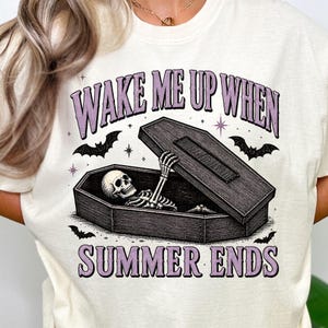 May include: Cream-colored t-shirt with a graphic featuring a skeleton rising from a coffin. The text "WAKE ME UP WHEN SUMMER ENDS" is in purple. Bats and stars surround the coffin, creating a spooky, Halloween-themed design.