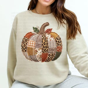 May include: A beige sweatshirt featuring a patchwork pumpkin design. The pumpkin is composed of various fabric patterns, including leopard print, floral, and plaid, in shades of orange, brown, and tan. The pumpkin has a brown stem and green leaves.