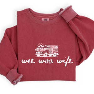 May include: A maroon-colored sweatshirt with a white graphic of a fire truck and the words "wee woo wife" in a cursive font. The sweatshirt has a crew neck and long sleeves with ribbed cuffs.