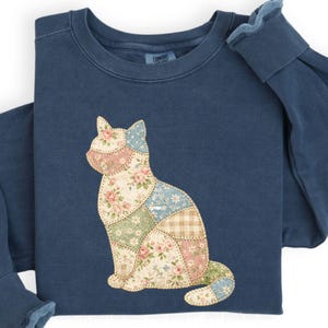 May include: A navy blue sweatshirt featuring a patchwork cat design. The cat is composed of various floral and patterned fabrics in shades of pink, blue, green, and tan. The sweatshirt has long sleeves and a crew neck.