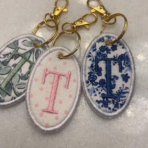 Może przedstawiać: Trzy haftowane breloczki do kluczy ze złotymi zapięciami. Każdy owalny breloczek ma literę "T" w różnych wzorach: kwiatowym, w kropki i niebieskim wzorze kwiatowym. Breloczki leżą na białej powierzchni.