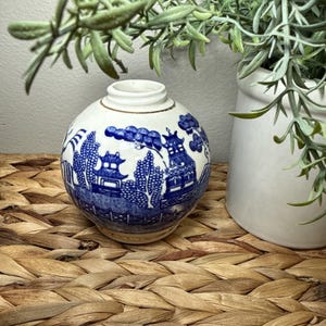 May include: A small, round, white and blue ceramic vase with a Willow pattern design. The vase features a blue landscape scene with pagodas and trees. The top rim has a gold accent. Decorative vase for home decor.