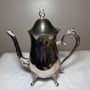 May include: A silver-colored teapot with a curved spout and ornate handle. The teapot has a lid with a decorative finial and rests on three small feet. The design features floral accents.