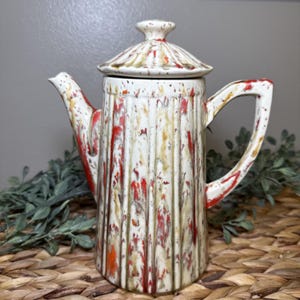 May include: A ceramic coffee pot featuring a vertical ribbed texture and a speckled glaze in shades of cream, bright red, and muted olive green. The pot includes a matching lid with a small knob handle, a curved spout, and a sturdy side handle.
