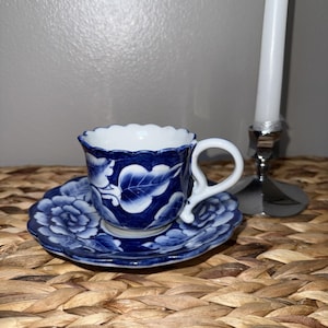 May include: A blue and white porcelain teacup and saucer set. The cup and saucer feature a floral design with a scalloped edge. The teacup has a white interior and a curved handle. The set is placed on a woven surface.