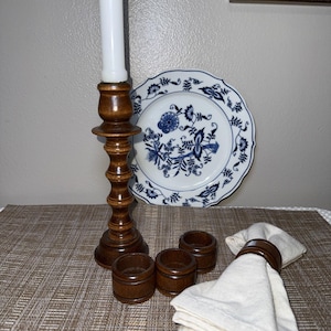 May include: A wooden candlestick with a white candle, a blue and white floral plate, three wooden napkin rings, and a folded white napkin. The items are arranged on a textured placemat.