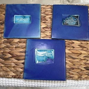 May include: Three square ceramic tiles with a deep blue glaze. Each tile features a central rectangular inset with a lighter blue background. The insets display raised relief designs of a grasshopper, a lizard, and a dragonfly in a metallic finish.