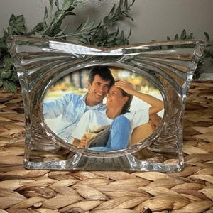 May include: A clear glass picture frame with a rectangular outer shape and a central oval opening. The frame features a textured, rippled surface design that radiates outward from the center. It holds a photograph of two people sitting together.