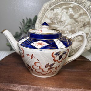 May include: A ceramic teapot featuring a cream-colored base with orange floral patterns and a deep blue geometric band around the top. The lid has a matching blue and orange design with gold trim accents on the handle, spout, and lid rim.