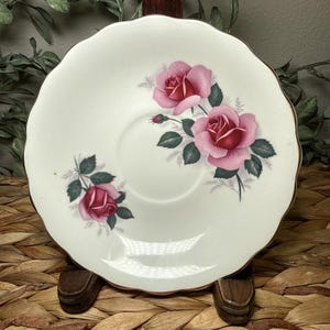 May include: A white porcelain saucer with a scalloped edge and a gold-trimmed rim. The surface features a floral pattern of three pink roses with dark green leaves and small grey sprigs arranged in two clusters on the off-white ceramic base.