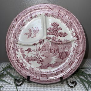 May include: A vintage divided plate with a red and white Willow pattern. The plate features three sections and a detailed landscape design with pagodas, bridges, and birds. The rim has a decorative border.