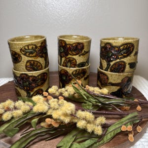 May include: Six ceramic cups with a floral design in shades of brown, green, and orange. The cups are stacked on a wooden tray, with decorative sprigs of yellow and green foliage.