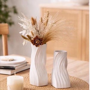 Peut inclure: Deux vases en céramique blanche avec un motif tourbillonnant texturé. L'un des vases contient des fleurs et du feuillage séchés. Les vases sont posés sur un set de table tissé sur une table en bois, avec une bougie allumée et des assiettes à proximité.