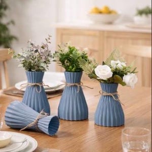 May include: A collection of blue vases with a pleated design, each holding greenery or white flowers. The vases are tied with twine and arranged on a wooden table, creating a decorative centerpiece. One vase is lying on its side.